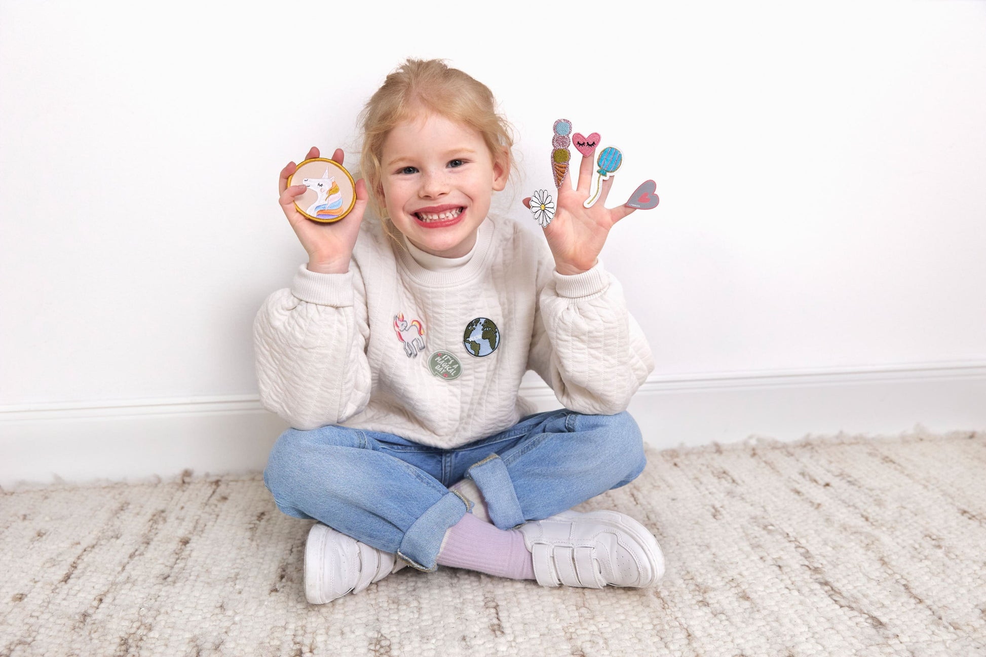 Lachendes Kind sitzt auf dem Boden und zeigt Laessig Patches & Anhaenger aus dem Einhorn Set, mit einem Einhorn-Patch in der linken Hand und verschiedenen Dekorationen in der rechten Hand.