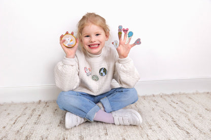 Lachendes Kind sitzt auf dem Boden und zeigt Laessig Patches & Anhaenger aus dem Einhorn Set, mit einem Einhorn-Patch in der linken Hand und verschiedenen Dekorationen in der rechten Hand.