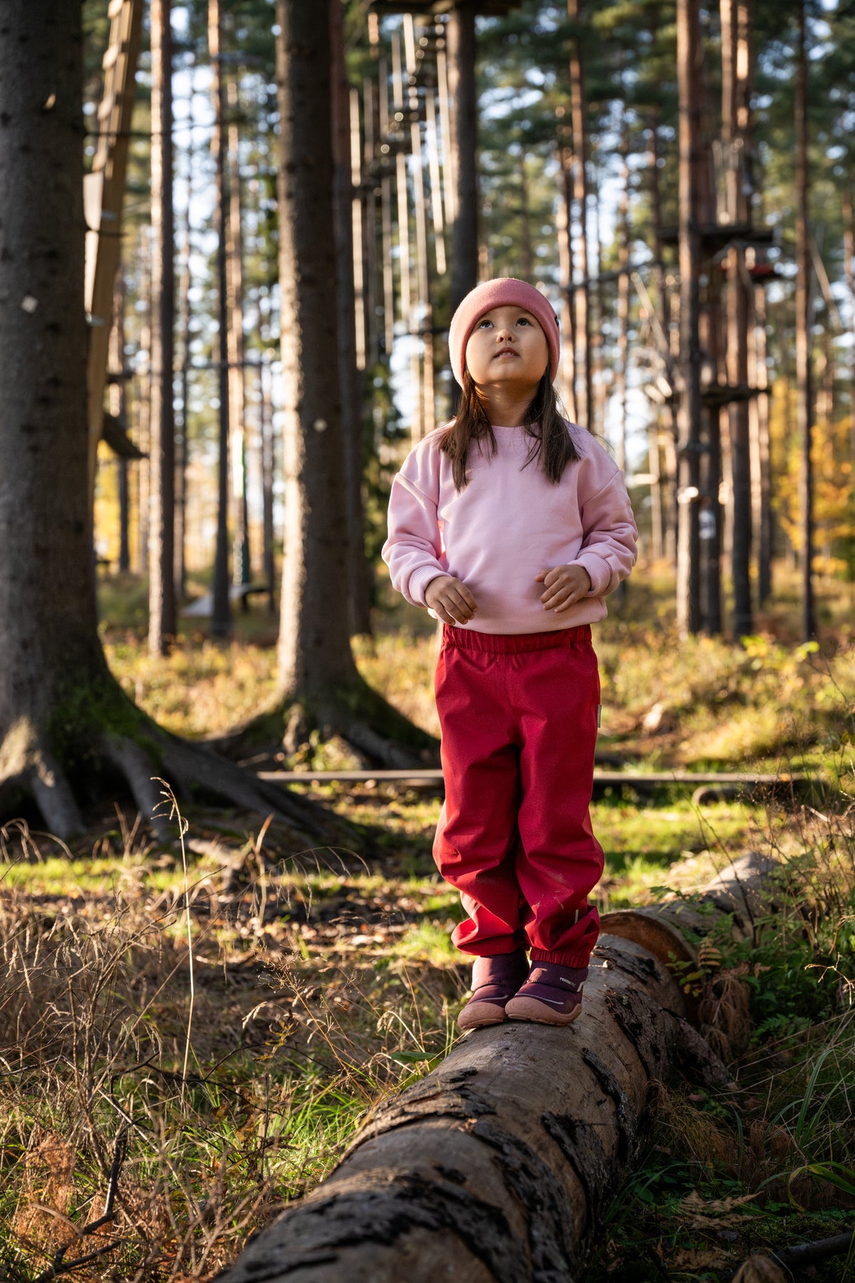 Kind steht mit der Reima Kaura Regenhose Cranberry auf einem Baumstamm im Wald bei Bärechind